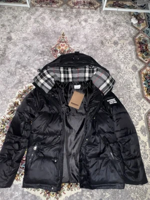 Svart pufferjacka från Burberry - Snygg svart pufferjacka från Burberry med klassiskt rutigt foder i huvan. Jackan har dragkedjor på fickorna, tryckknappar och en logga på ärmen. Perfekt för kalla vinterdagar och riktigt trendig look, storlek M men passar mer som S