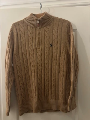 Beige halvzip stickad tröja Polo Ralph Lauren - Kabelstickad beige tröja från Polo Ralph Lauren med halv dragkedja och hög krage. Tröjan har lång ärm, ribbstickade muddar och den klassiska broderade loggan på bröstet. Perfekt för dig som gillar stilren och tidlös design.