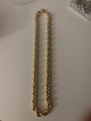 Cordell halsband(Guld 10mm 60cm) - Säljer ett snyggt guldfärgat halsband med tvinnad kedja. Halsbandet har en chunky och trendig design som verkligen sticker ut. Perfekt accessoar för att lyfta din outfit med en touch av guld. Materialet är metall och låset är av klassisk modell.