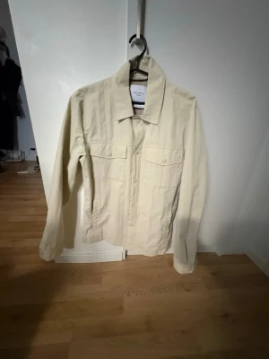 Beige overshirt från Les Deux - Stilren beige overshirt från Les Deux med klassisk krage och två bröstfickor med lock. Tillverkad i ett strukturerat bomullstyg som ger en avslappnad men snygg look. Perfekt att bära öppen över en t-shirt eller stängd som lätt jacka.