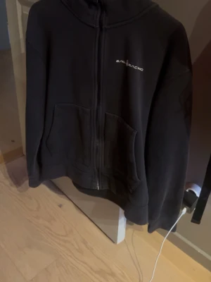 Svart hoodie från Sail Racing - Svart hoodie med dragkedja från Sail Racing. Klassisk modell med huva, två stora fickor framtill och diskret logga på bröstet. Tillverkad i mjukt material som känns skönt mot huden. Perfekt för en avslappnad och sportig stil.