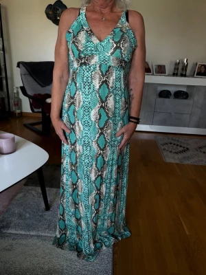 Grön långklänning med ormmönster - Säljer en snygg långklänning i grönt, beige och svart med ormmönster. Klänningen har smala axelband som korsas i ryggen och knyts baktill. V-ringad framtill och i ett mjukt, följsamt material. Perfekt för dig som vill sticka ut!