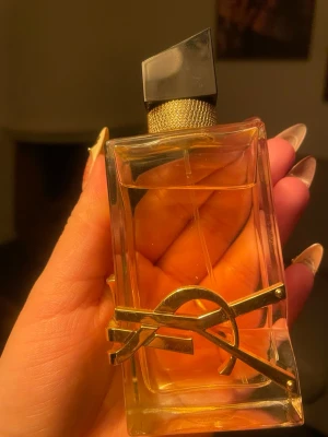 Yves Saint Laurent Libre EDT 50ml - Snygg parfymflaska från Yves Saint Laurent, modell Libre. Flaskan är rektangulär i genomskinligt glas med guldig detalj och svart asymmetrisk kork. Innehållet är bärnstensfärgat. Perfekt för dig som gillar lyxiga dofter och stilren design.
