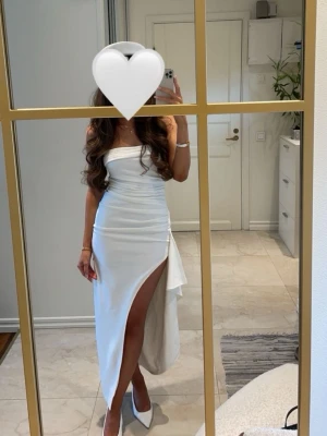 Vit långklänning med slits från SHOWPO - Elegant vit långklänning med axelbandslös design och hög slits på ena sidan. Klänningen har en figurnära passform och ett stilrent, minimalistiskt uttryck. Perfekt för dig som vill sticka ut med en klassisk och sofistikerad look.