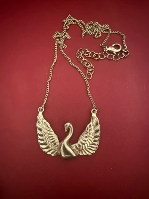 Silverfärgat halsband med svanmotiv - Unikt halsband i silverfärgad metall med en detaljrik svan med utbredda vingar som hänge. Kedjan har justerbar längd och ett klassiskt lås. Perfekt för dig som vill sticka ut med ett djurinspirerat smycke.