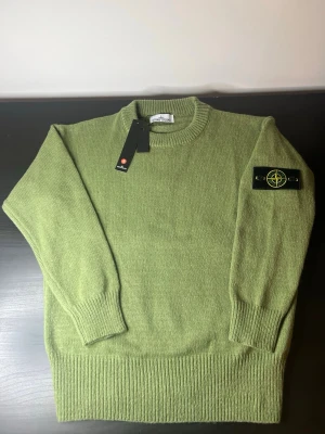 Grön stickad tröja från Stone Island - Säljer en snygg grön stickad tröja från Stone Island med klassisk rund hals och ribbade muddar. Tröjan har den ikoniska Stone Island-loggan på vänster ärm och är tillverkad i mjukt ullmaterial. Perfekt för dig som gillar stilrena plagg med exklusiv känsla.