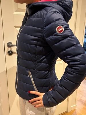 Mörkblå pufferjacka från Colmar - Snygg mörkblå dunjacka från Colmar med huva och dragkedja framtill. Jackan har två snedställda fickor med dragkedja och ett broderat Colmar-märke på ärmen. Fodret har en kontrasterande rosa färg. Perfekt för kalla vinterdagar.