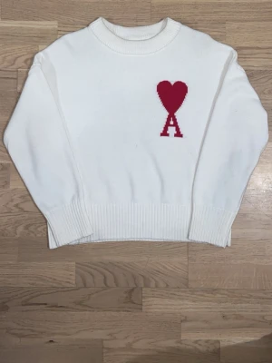 Ami Paris - Vit stickad tröja från Ami Paris med röd hjärtapplikation och bokstaven A på bröstet. Tröjan har ribbade muddar vid hals, ärmslut och nederkant samt en klassisk rund halsringning. Perfekt för dig som gillar stilrena detaljer och vill sticka ut med en ikonisk design. Jag har använt den några gånger så prislappen är inte kvar så den är inte helt ny skick.