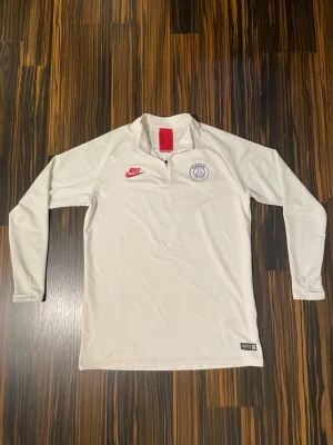 Vit långärmad Nike PSG träningströja - Säljer en vit långärmad träningströja från Nike med PSG-logga på bröstet och röd Nike Swoosh. Tröjan har halv dragkedja framtill och är tillverkad i ett lätt, tekniskt material som passar perfekt för träning eller uppvärmning.