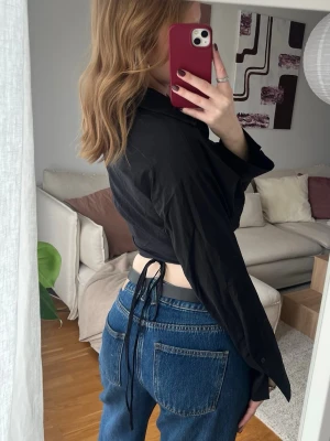 Svart skjorta/topp med snörning - Säljer en svart croppad skjorttopp med långa ärmar och snörning i ryggen. Toppen har krage och v-ringning framtill, vilket ger en edgy och trendig look. Perfekt att styla med höga jeans eller kjol.