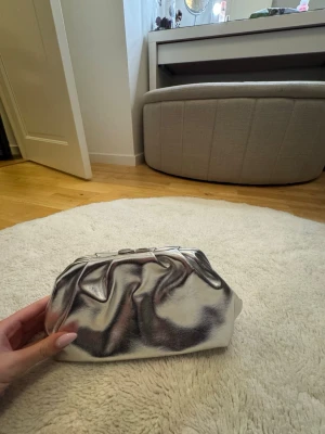 Silvrig kuvertväska i metallic - Säljer en snygg silvrig kuvertväska i metallic-look med mjuk, rynkad design. Väskan har en rektangulär botten och stängs upptill med metallram. Perfekt för dig som vill sticka ut med en trendig accessoar.