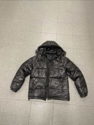 Svart pufferjacka från Polo Ralph Lauren - Säljer en svart pufferjacka från Polo Ralph Lauren med huva och dragkedja. flat DNA-logga i rött på bröstet och två sidofickor med dragkedja. Jackan är vadderad och har en glansig finish, perfekt för kalla vinterdagar.