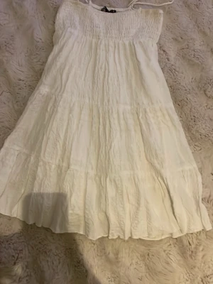 Vit sommarklänning från Zara - Luftig vit klänning från Zara med smock upptill och tunna axelband. Klänningen har en flowy passform och volangdetaljer nedtill, perfekt för varma dagar. Tillverkad i mjukt bomullstyg som känns skönt mot huden.