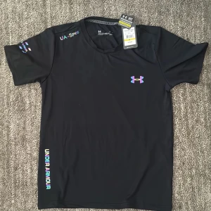 Svart t-shirt från Under Armour - Svart sport-t-shirt från Under Armour med färgglada loggor och tryck på bröstet, ärmen och längs sidan. Tillverkad i ett lätt och snabbtorkande material, perfekt för träning. Rund halsringning och korta ärmar ger en klassisk sportig look.                           Strl: M 
