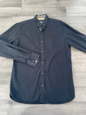 Svart långärmad skjorta med knappar - Stilren svart skjorta med lång ärm och klassisk krage. Skjortan har knäppning framtill med matchande svarta knappar och är tillverkad i ett mjukt bomullstyg. Perfekt för en clean och enkel look.