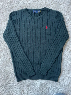 Mörkgrön stickad tröja Polo Ralph Lauren - Mörkgrön kabelstickad tröja från Polo Ralph Lauren med rund halsringning och röd broderad logga på bröstet. Tröjan har långa ärmar och ribbade muddar vid ärmslut och nederkant.Perfekt för dig som gillar klassisk och stickat