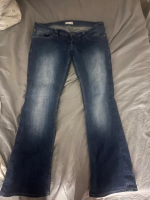 Blå bootcut jeans från Gina Tricot. - Snygga blå bootcut jeans från Gina med klassisk femficksdesign och ljusa slitningar framtill. Jeansen har en läderpatch bak i midjan och dekorativa sömmar på bakfickorna. Perfekta för en avslappnad och trendig look.