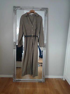 Beige långklänning i linne med bälte - Stilren beige långklänning med knappar framtill och klassisk krage. Klänningen har långa ärmar, två bröstfickor och ett svart bälte i midjan som ger en snygg siluett. Perfekt för dig som gillar en enkel men trendig look.