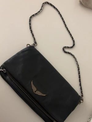 Svart axelväska från Zadig & Voltaire - Cool svart axelväska i läder från Zadig & Voltaire med silverfärgad kedjerem och ikonisk vinge i metall framtill. Väskan har dragkedja och en stilren, avlång form som passar perfekt till din outfit.