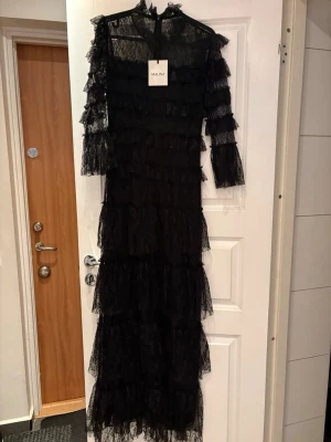 Svart lång spetsklänning från Malina - Svart långklänning från Malina i spets med volangdetaljer och trekvartsärm. Klänningen har transparenta partier och en elegant, figurnära siluett. Perfekt för dig som vill sticka ut med en dramatisk look.