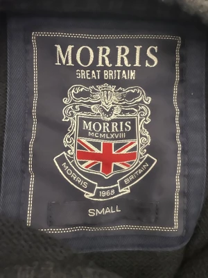 Mörkgrå stickad tröja från Morris - Snygg mörkgrå stickad tröja från Morris med broderad logga och brittisk flagga på bröstet. Tröjan har rund halsringning och ribbade muddar vid ärmslut och nederkant. Perfekt för dig som gillar klassisk brittisk stil.