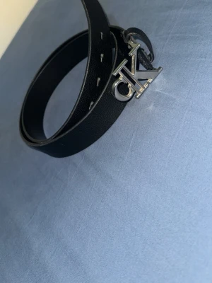 Svart skärp från Calvin Klein - Stilrent svart skärp från Calvin Klein med ett snyggt CK-spänne i silverfärg. Skärpet är gjort i skinn och passar perfekt till jeans eller kostymbyxor. Enkel design med flera hål för justerbar passform.