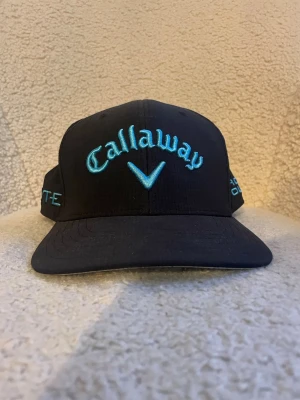Svart Callaway keps med blå brodyr - Snygg svart keps från Callaway med broderad logga och text i ljusblått framtill och på sidorna. Modellen har böjd skärm och justerbar passform. Perfekt accessoar för golfbanan eller sportiga dagar.