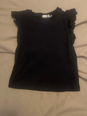 Svart topp med volang från Uniq Studio - Snygg svart topp från Uniq Studio med volangdetaljer vid axlarna. Modellen är ärmlös och har en rund halsringning. Perfekt att matcha med jeans eller kjol för en trendig look.