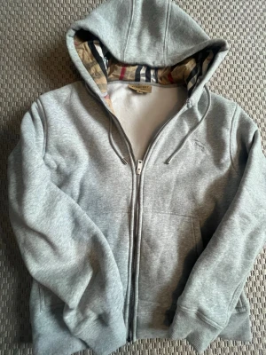 Grå hoodie från Burberry med dragkedja - Snygg grå hoodie från Burberry med dragkedja och klassiskt rutigt foder i huvan. Broderad logga på bröstet och ribbade muddar. Perfekt för en chill och stilren look. Storlek smal lite större 