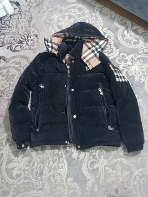 Svart pufferjacka från Burberry - Säljer en svart pufferjacka från Burberry med klassiskt rutigt foder och huva. Jackan har dragkedjor och tryckknappar framtill, samt två snedställda fickor med dragkedja. Ikoniskt Burberry-märke på bröstet och rutiga detaljer på axel och insida.