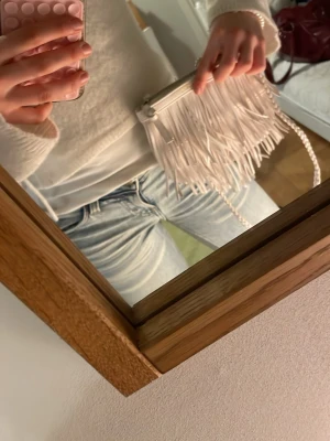 Vit kuvertväska med fransar - Säljer en superfin vit kuvertväska från Rebecca Minkoff med långa fransar framtill och flätad axelrem. Väskan är gjord i läder och har guldfärgad logga upptill. Perfekt accessoar för att lyfta din outfit!