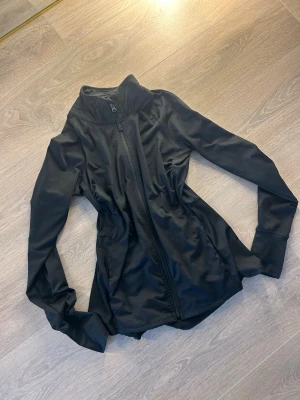 Svart träningsjacka med dragkedja - Snygg svart träningsjacka med hel dragkedja framtill och hög krage. Tillverkad i ett stretchigt och lätt material som passar perfekt för löpning eller gym. Jackan har långa ärmar och en smal passform som ger en sportig look.