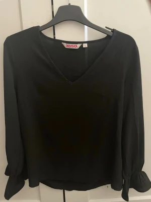 Svart blus med V-ringning från Indiska - Stilren svart blus från Indiska med V-ringad hals och långa ärmar med knyt vid ärmsluten. Blusen har en rak passform och är tillverkad i ett lätt, flowy tyg som ger en snygg siluett.