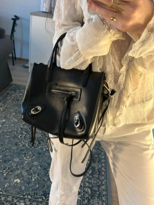 Svart handväska med silverdetaljer - Snygg svart handväska i slätt konstläder med både handtag och avtagbar axelrem. Väskan har en dragkedjeficka framtill och coola silverfärgade spännen som detaljer. Perfekt storlek för det viktigaste och ger en edgy vibe till din outfit.