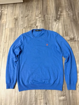 Blå tröja från Sail Racing - Snygg blå tröja från Sail Racing med rund halsringning och diskret orange logga broderad på bröstet. Tröjan har ribbade muddar vid ärmslut och nederkant. Perfekt till jeans eller chinos för en clean look.