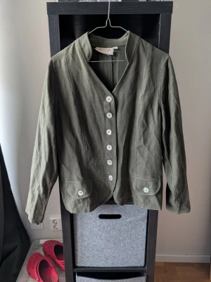 Olivgrön kavaj från Anna - Stilren olivgrön kavaj från Linna med v-ringad krage och sex vita knappar framtill. Två fickor med knappdetaljer och rak passform. Tillverkad i ett tyg som ser ut att vara linne eller bomull, perfekt för en avslappnad men snygg look.