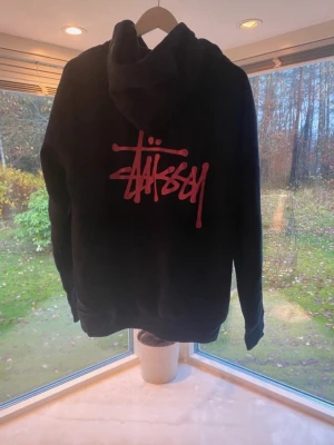 Svart Stüssy hoodie med rosa logga - Säljer en svart hoodie från Stüssy med den ikoniska loggan i risa  tryck både på bröstet och stort på ryggen. Hoodien har huva med dragsko och en klassisk magficka. Overzise hoodie. Perfekt för dig som gillar streetwear och vill ha en stilren men ändå fet look. L men passar som M 