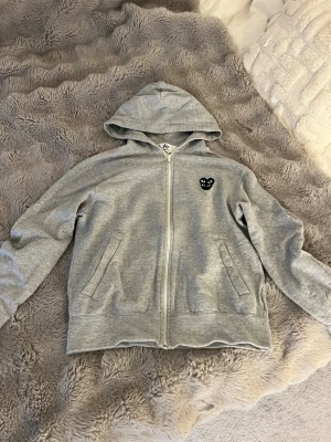 Grå hoodie från Comme des Garçons Play - Säljer en grå zip-hoodie från Comme des Garçons Play med det ikoniska svarta hjärta-logotypen på bröstet. Hoodien har dragkedja, två fickor fram och en klassisk huva. 