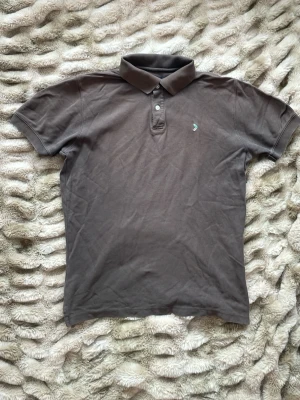 Brun pikétröja från Boomerang - Klassisk brun pikétröja från Boomerang med korta ärmar och krage. Tröjan har två knappar vid halsen och en liten broderad logga på bröstet. Tillverkad i mjuk bomull som känns skön mot huden. Pikén har ett litet hål under armen som knappast syns. Har även en liten fläck på axeln 