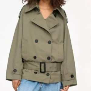 Olivgrön trenchcoat med bälte - Stilren olivgrön trenchcoat med dubbelknäppning och brett bälte i midjan. Jackan har klassisk krage, axelklaffar och svarta knappar. Passar perfekt till jeans för en avslappnad men trendig look.