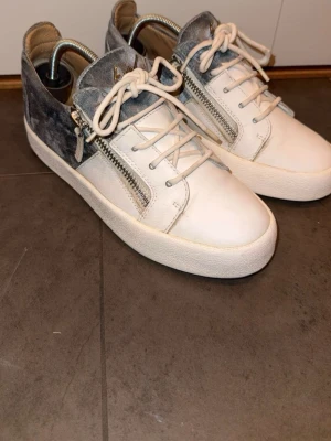 Vita och gråa sneakers med dragkedja - Snygga sneakers från Giuseppe Zanotti i vitt och grått läder med silvriga dragkedjor på sidorna. Skorna har vita snören, tjock vit sula och en cool kontrast mellan den ljusa framsidan och den mörkare baksidan. Perfekta för dig som gillar streetstyle och unika detaljer.