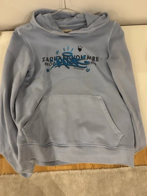 Ljusblå hoodie Zadig & Voltaire - Ljusblå hoodie från Zadig & Voltaire med coolt blått och svart tryck framtill. Stor känguruficka och huva. Skön och avslappnad passform, perfekt för en chill look. Materialet känns mjukt och bekvämt mot huden.