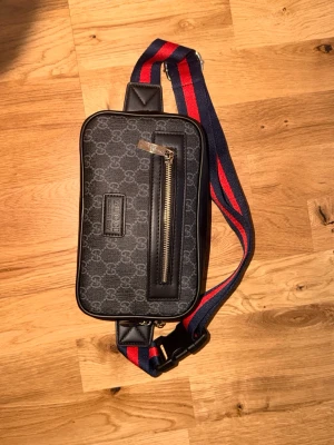 Gucci axelväska med monogram - Snygg svart axelväska från Gucci med klassiskt GG-monogram och en framficka med dragkedja. Väskan har en bred axelrem i blått och rött, samt ett rymligt innerfack med extra ficka. Tillverkad i canvas och läderdetaljer.