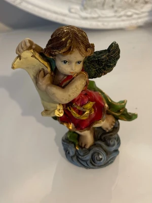 Färgglad ängel i keramik - Dekorativ ängelfigur i keramik med gröna vingar, röd klänning och guldfärgade detaljer. Ängeln sitter på ett blått moln och håller i en guldfärgad rulle. Perfekt som prydnad eller samlarobjekt.