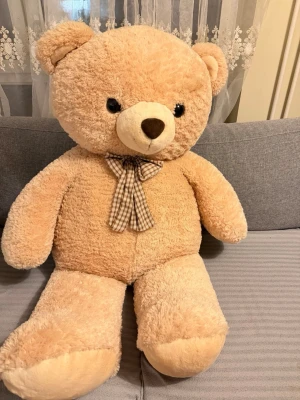 Stor beige nallebjörn med rosett - Mysig och stor nallebjörn i beige plysch med svartbruna ögon och nos. Nallen har en brun- och vitrutig rosett runt halsen och är perfekt som dekoration eller gosedjur. Tillverkad i mjukt material och har klassisk rund form.