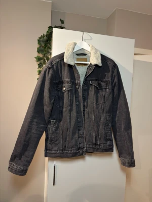 Svart jeansjacka med teddykrage - Säljer en svart jeansjacka från Levi's med vit teddykrage och teddyfoder. Klassisk modell med två bröstfickor med knapp och två sidofickor. Jackan har knappstängning framtill och detaljerade sömmar. Perfekt för dig som vill ha en cool och tidlös look.