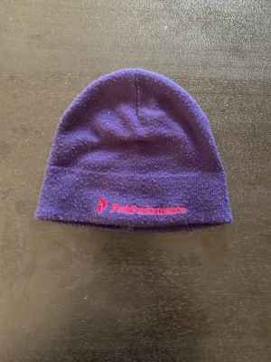 Lila mössa från Peak Performance - Säljer en lila mössa från Peak Performance med broderad logga i rosa framtill. Klassisk modell i mjukt stickat material som sitter skönt på huvudet. Perfekt accessoar för kyliga dagar och enkel att matcha med olika outfits.