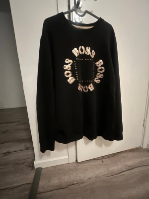 Svart sweatshirt från Hugo Boss - Cool svart sweatshirt från Hugo Boss med rund hals och tryck i vitt och beige där det står 'BOSS' i cirkel på bröstet. Tröjan har en avslappnad passform och långa ärmar, perfekt för en casual och trendig look.
