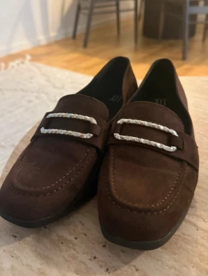 Bruna mockaloafers med silverdetalj - Snygga loafers i mörkbrun mocka med dekorativa silverfärgade detaljer över vristen. Skorna har klassisk, stilren design med låg klack och rundad tå. Perfekta för dig som gillar en tidlös look med en twist.