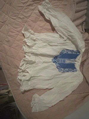 Vit blus med blå brodyr Les Jumelles - Supersöt vit blus från Les Jumelles med långärmad design och blå broderade detaljer framtill. Blusen har volang vid ärmslut och hals, samt en luftig och bohemisk känsla. Perfekt för dig som gillar unika detaljer och romantisk stil. Passar ungefär M. 